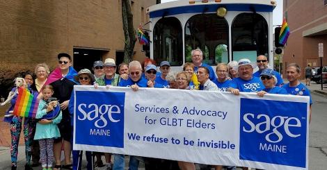 A photo of many SAGE Maine members at a pride parade. They are holding a banner with the SAGE Maine logo.